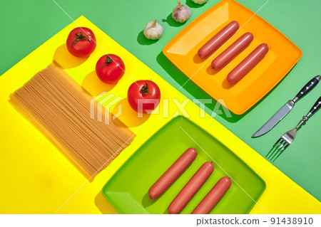 A lot of raw sausages on plate. On green and yellow background with pasta and vegetables, top view. 91438910