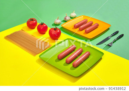 A lot of raw sausages on plate. On green and yellow background with pasta and vegetables, top view. A lot of raw sausages on plate. On green and yellow background with pasta and vegetables, top view. 91438912