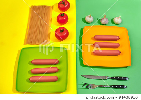 A lot of raw sausages on plate. On green and yellow background with pasta and vegetables, top view. 91438916