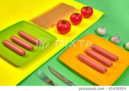 A lot of raw sausages on plate. On green and yellow background with pasta and vegetables, top view. A lot of raw sausages on plate. On green and yellow background with pasta and vegetables, top view. 91438924