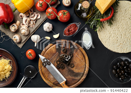Cutting wooden board with traditional pizza preparation ingredients: cheese, tomatoes sauce, olives, olive oil, pepper, spices. Black texture table background 91440422