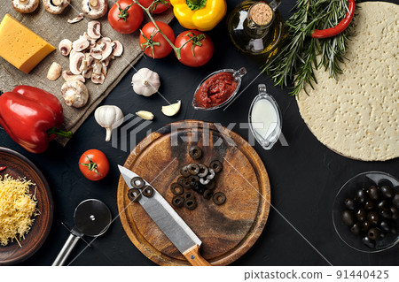 Cutting wooden board with traditional pizza preparation ingredients: cheese, tomatoes sauce, olives, olive oil, pepper, spices. Black texture table background 91440425