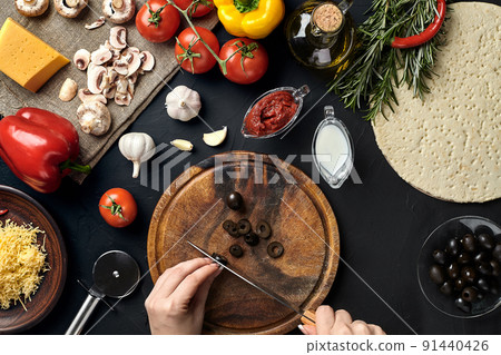 Female hand cut olives on wooden board on kitchen table, around lie ingredients for pizza: vegetables, cheese and spices. 91440426