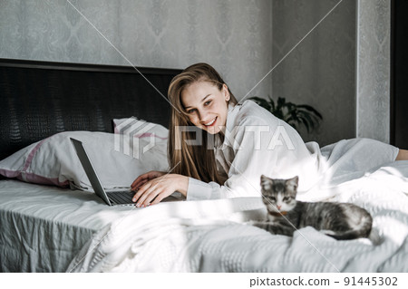 Work from home, remote online work, home office. Woman working on laptop in bed and playing with cute kitten Work from home, remote online work, home office. Woman working on laptop in bed and playing with cute kitten 91445302