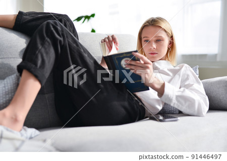 Woman lies on couch and reads book. 91446497