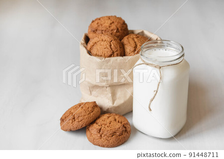 Milk in a glass jar and oatmeal cookies in a paper bag on a white table background. Copy, empty space for text Milk in a glass jar and oatmeal cookies in a paper bag on a white table background. Copy, empty space for text 91448711