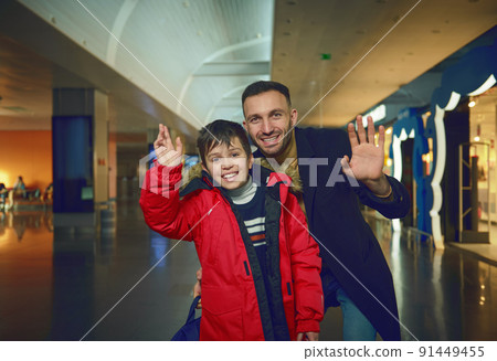 Handsome multi-ethnic man- dad and his son waving looking at camera while standing outside duty free shops in international airport departure terminal Handsome multi-ethnic man- dad and his son waving looking at camera while standing outside duty free shops in international airport departure terminal 91449455