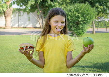 A teenage girl chooses between a hamburger and fruit outdoors. 91449539