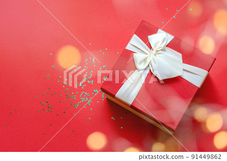 Defocused round lights in the foreground. Beautiful red gift box with satin light ribbon on red background. Green stars shine on the table. Gift for Christmas and New Year. Copy space 91449862