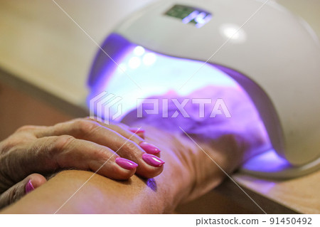 Woman hand inside UV LED lamp for drying pink nails with gel method, plate curing close up. Manicure process. Curing recently applied gel nail polish or hybrid varnish in beauty salon, beautician. Woman hand inside UV LED lamp for drying pink nails with gel method, plate curing close up. Manicure process. Curing recently applied gel nail polish or hybrid varnish in beauty salon, beautician. 91450492