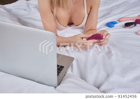 A woman lies in bed holding a clitoral vibrator and watching porn on a laptop. The girl has sex online 91452456