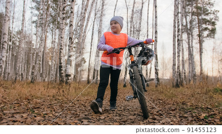 One caucasian children walk with bike in autumn park. Little girl walking black orange cycle in forest. Kid goes do bicycle sports. Biker motion ride with backpack and helmet. Mountain bike hard tail. 91455123