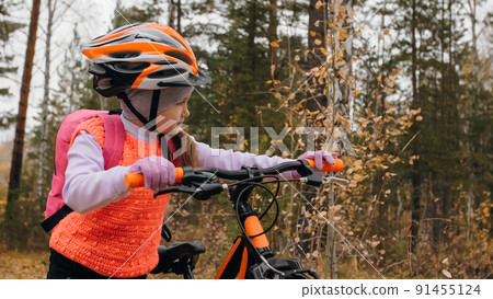 One caucasian children walk with bike in autumn park. Little girl walking black orange cycle in forest. Kid goes do bicycle sports. Biker motion ride with backpack and helmet. Mountain bike hard tail. 91455124
