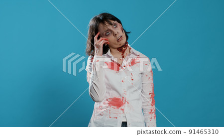 Brain-eating spooky zombie woman having an idea on blue background. Smart pensive looking crazy monster with bloody and deep wounds thinking about something while looking at camera. Studio shoot 91465310