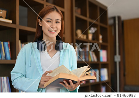 Attractive young Asian female college student standing in the library with a book. 91466988
