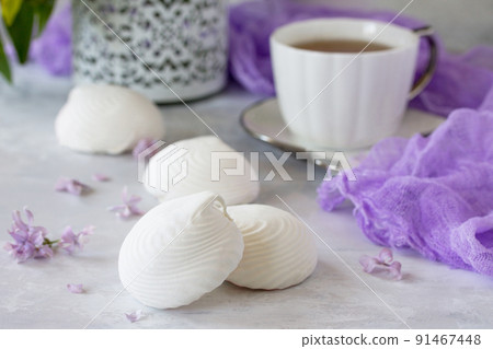 Delicate vanilla marshmallow on a white delicate plate. Still life with marshmallows, a cup of tea and lilac lilac on a background of ancient stone. Delicate vanilla marshmallow on a white delicate plate. Still life with marshmallows, a cup of tea and lilac lilac on a background of ancient stone. 91467448