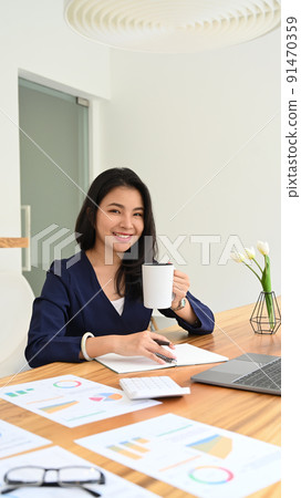 Pleased asian business entrepreneur drinking coffee and working with financial document at office desk Pleased asian business entrepreneur drinking coffee and working with financial document at office desk 91470359