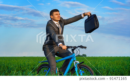 businessman rides a bicycle through a green grass field, dressed in a business suit, he has a briefcase and documents, beautiful nature in spring, business concept 91471165