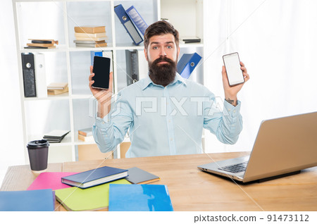 Serious businessman holding mobile phones in hands sitting at office desk, choice Serious businessman holding mobile phones in hands sitting at office desk, choice 91473112