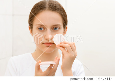 face of a teenage girl, she looks at herself in the mirror and takes care of her skin, does cosmetic procedures using a cotton pad, good and clean skin is a concept of beauty and health face of a teenage girl, she looks at herself in the mirror and takes care of her skin, does cosmetic procedures using a cotton pad, good and clean skin is a concept of beauty and health 91473118