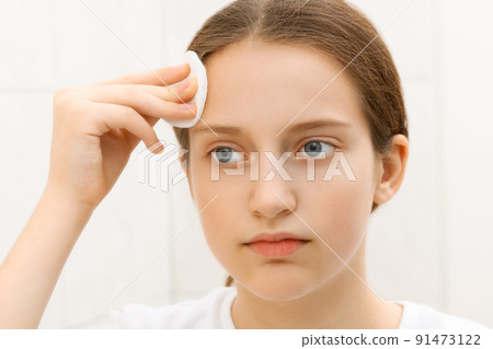 face of a teenage girl, she looks at herself in the mirror and takes care of her skin, does cosmetic procedures using a cotton pad, good and clean skin is a concept of beauty and health 91473122