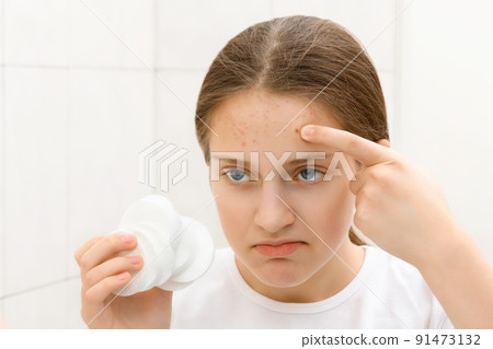 face of a teenage girl with pimples, she looks at herself in the mirror and takes care of her skin, does cosmetic procedures using a cotton pad face of a teenage girl with pimples, she looks at herself in the mirror and takes care of her skin, does cosmetic procedures using a cotton pad 91473132
