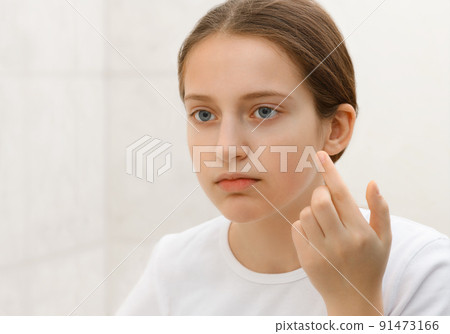 the face of a teenage girl, she looks at herself in the mirror and touches her face with her hand, good and clean skin is a concept of beauty and health the face of a teenage girl, she looks at herself in the mirror and touches her face with her hand, good and clean skin is a concept of beauty and health 91473166