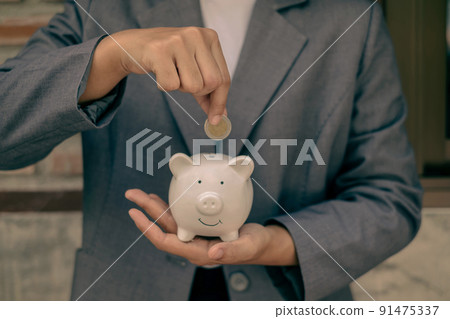 Asian businesswomen hold piggy banks and put coins in piggy banks to save money with coins to step into business, grow successful, and save money. savings concept Asian businesswomen hold piggy banks and put coins in piggy banks to save money with coins to step into business, grow successful, and save money. savings concept 91475337