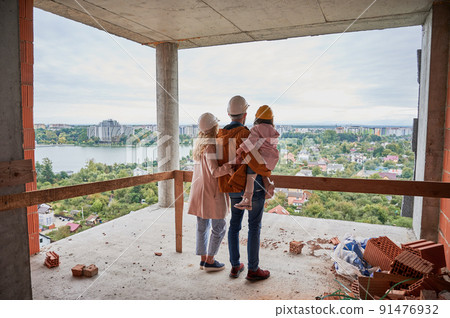 Back view of parents with daughter standing inside apartment building under construction. Man and woman with child wearing building helmets while enjoying city view from future home. 91476932