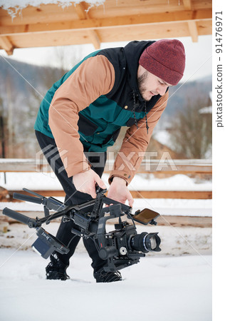 Videographer man walking on snow, shooting footage, using camera mounted on gimbal stabilizer equipment in winter. 91476971
