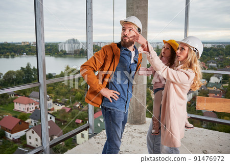 Woman holding daughter and pointing at something while standing next to husband at construction site. Family with child future homeowners observing apartment building under construction. Woman holding daughter and pointing at something while standing next to husband at construction site. Family with child future homeowners observing apartment building under construction. 91476972