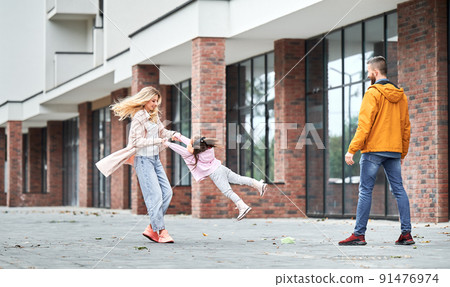 Full length of smiling blonde woman spinning daughter holding kid hands while spending time with family outdoors. Loving parents having fun with child on the street outside new building. 91476974