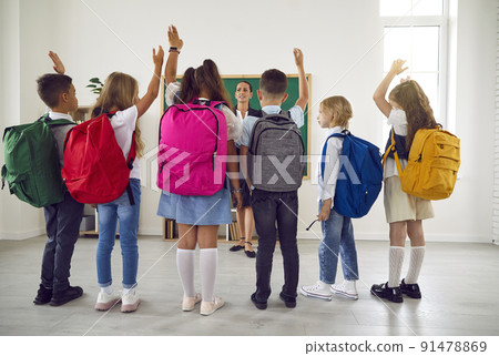 Children with large backpacks on their shoulders raise their hands talking with teacher in classroom 91478869