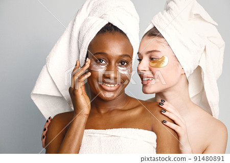 Two young women in white bathrobes standing isolated on white. Girls wearing white towels on their hair. Girls have a eye patches on their faces. 91480891