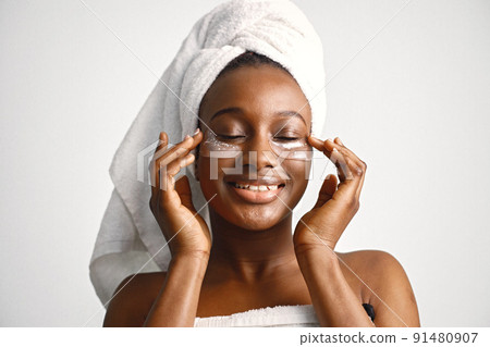 Black young woman standing isolated on white background with patches on her face. Girl has her hair wrapped in a towel. Black woman enjoy after body treatment. Black young woman standing isolated on white background with patches on her face. Girl has her hair wrapped in a towel. Black woman enjoy after body treatment. 91480907