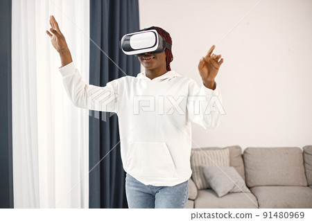 Black young woman standing near coach wearing virtual reality glasses. Black girl wearing white hoodie and jeans. Girl spending time alone. 91480969