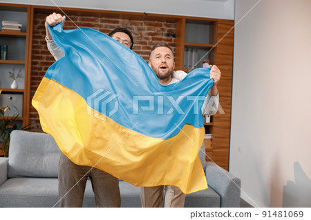 Male friends gathered at home to watch a football match. Two men standing and holding a big flag of Ukraine in hands. Men actively support their favorite team and feeling satisfied. Male friends gathered at home to watch a football match. Two men standing and holding a big flag of Ukraine in hands. Men actively support their favorite team and feeling satisfied. 91481069