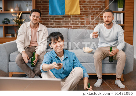 Male friends gathered at home to watch a football match. Asian man in a middle has a TV remote control. Men actively support their favorite team and feeling disapointed. Male friends gathered at home to watch a football match. Asian man in a middle has a TV remote control. Men actively support their favorite team and feeling disapointed. 91481090
