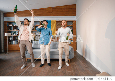 Male friends gathered at home to watch a football match. Men standing in front of big flag of Ukraine on a wall. Men actively support their favorite team and feeling satisfied. Male friends gathered at home to watch a football match. Men standing in front of big flag of Ukraine on a wall. Men actively support their favorite team and feeling satisfied. 91481109