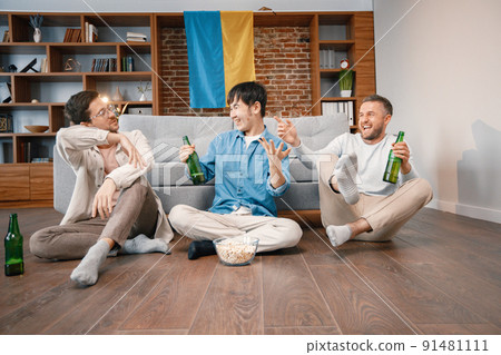 Male friends gathered at home to watch a football match. Men sitting on the couch in front of big flag of Ukraine on a wall. Men actively support their favorite team and feeling satisfied. Male friends gathered at home to watch a football match. Men sitting on the couch in front of big flag of Ukraine on a wall. Men actively support their favorite team and feeling satisfied. 91481111