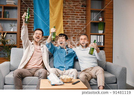 Male friends gathered at home to watch a football match. Men sitting on the couch in front of big flag of Ukraine on a wall. Men actively support their favorite team and feeling satisfied. 91481119