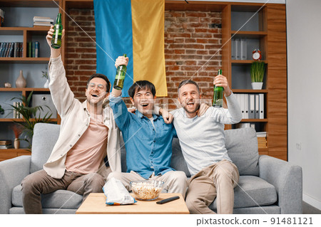 Male friends gathered at home to watch a football match. Men sitting on the couch in front of big flag of Ukraine on a wall. Men actively support their favorite team and feeling satisfied. 91481121