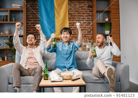 Male friends gathered at home to watch a football match. Men sitting on the couch in front of big flag of Ukraine on a wall. Men actively support their favorite team and feeling satisfied. 91481123