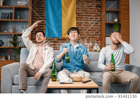 Male friends gathered at home to watch a football match. Men sitting on the couch in front of big flag of Ukraine on a wall. Men actively support their favorite team and feeling disapointed. 91481130