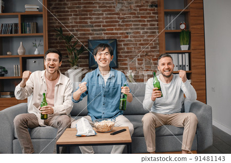 Front view of male friends gathered at home to watch a football match. Men sitting on the couch in front of the big screen TV and eating snacks. Men actively support their favorite team and feeling Front view of male friends gathered at home to watch a football match. Men sitting on the couch in front of the big screen TV and eating snacks. Men actively support their favorite team and feeling 91481143