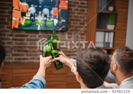 Focus on a bottles of beer. Back view of male friends gathered at home to watch a football match. Men actively support their favorite team and comment on the match 91481159