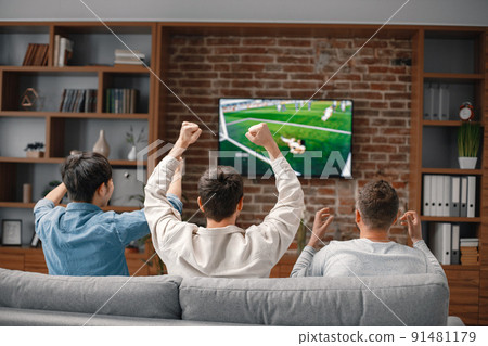 Back view of male friends gathered at home to watch a football match. Men sitting on the couch in front of the big screen TV. Men actively support their favorite team and comment on the match Back view of male friends gathered at home to watch a football match. Men sitting on the couch in front of the big screen TV. Men actively support their favorite team and comment on the match 91481179