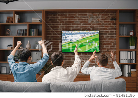 Back view of male friends gathered at home to watch a football match. Men sitting on the couch in front of the big screen TV. Men actively support their favorite team and comment on the match 91481190