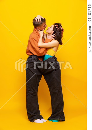 Creative colorful portrait of man and woman in bright clothes, standing together in one pants isolated over yellow background. 91481716