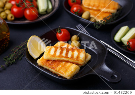 Grilled halloumi cheese in a black pan with olives, tomatoes, cucumbers, thyme and lemon. Traditional cheese of the Eastern Mediterranean. Black background, selective focus, close-up. 91482269